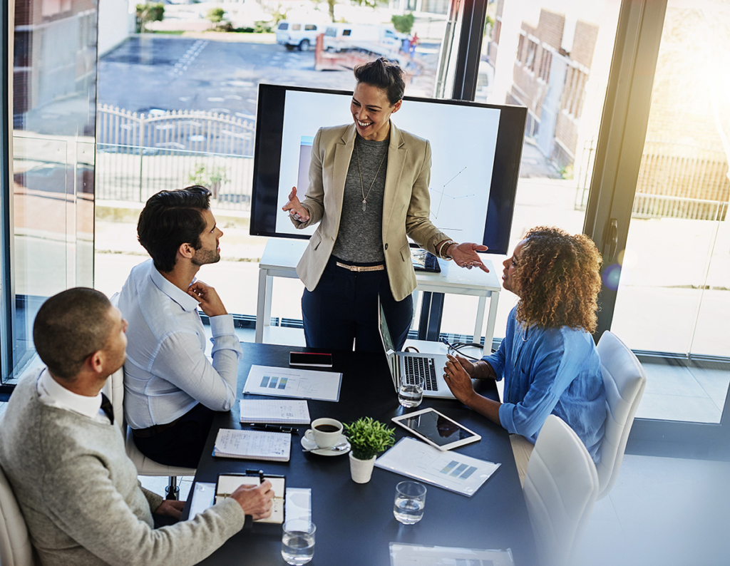 A bright, glassโwalled conference room: a smiling woman in a beige blazer stands at the head of a black table, gesturing as she leads a presentation displayed on a large monitor behind her. Three colleaguesโa man in a gray sweater taking notes, a man in a white shirt listening thoughtfully, and a woman in a blue blouse seated at a laptopโsit around the table strewn with notebooks, a tablet, coffee cups, and glasses of water. Through the floorโtoโceiling windows, a parking lot and city buildings are visible.