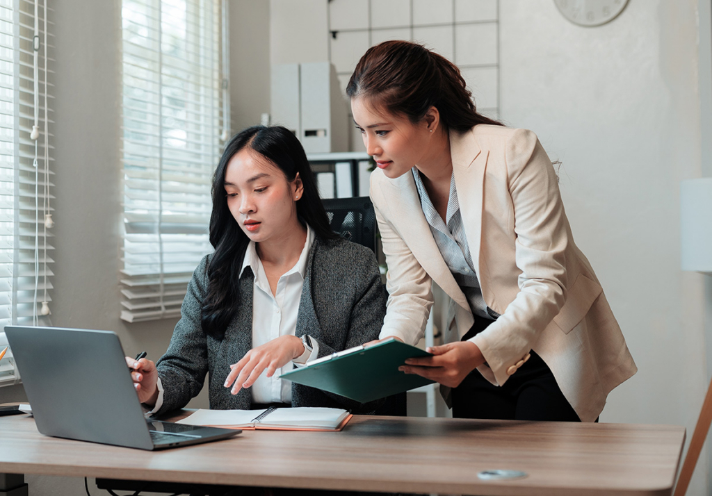 Two businesswomen collaborating at a desk in a modern office, one seated using a laptop and pointing at documents while the other stands beside her holding a clipboard and reviewing the work together.